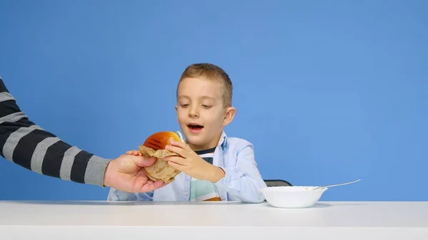 Мальчик сидит за столом и не хочет есть кашу на синем фоне. Мальчику дают гамбургер, и он радуется. Понятие здоровой и нездоровой пищи. Фаст-фуд — стоковое фото