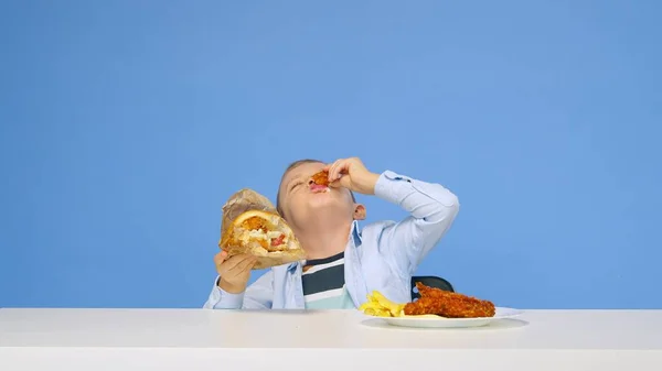 A fiú az asztalnál ül, gyorskaját eszik, és kék háttérrel örül. Az egészséges és egészségtelen ételek fogalma. Gyorsétterem — Stock Fotó