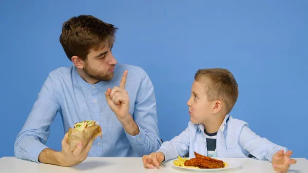 Ein junger Mann und ein Junge sitzen am Tisch, der Mann erlaubt dem Jungen nicht, Fast Food zu essen. Das Konzept der gesunden und ungesunden Ernährung. Fast food — Stockfoto