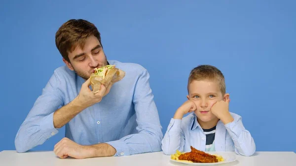 Egy fiatalember és egy fiú ül az asztalnál, a férfi nem engedi a fiúnak, hogy gyorskaját egyen. Az egészséges és egészségtelen ételek fogalma. Gyorsétterem — Stock Fotó