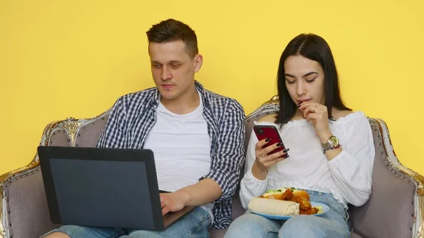 El chico y la chica comen comida rápida, el chico está sentado en un portátil, y la chica está mirando el teléfono en un fondo amarillo. El concepto de comida saludable y poco saludable. Comida rápida — Foto de Stock