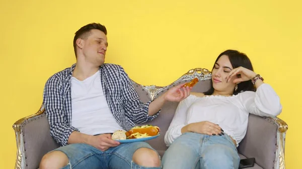 Chlápek jí rychlé občerstvení, sedí na gauči, a dívka drží dietu, na žlutém pozadí. pár dívající se do kamery. Koncept zdravé a nezdravé stravy. fast food — Stock fotografie