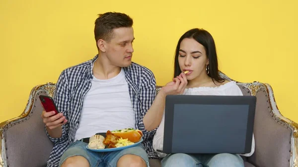 De man en het meisje eten fastfood, het meisje zit achter een laptop, en de man kijkt naar de telefoon, op een gele achtergrond. Het concept van gezond en ongezond voedsel. Fast food — Stockfoto
