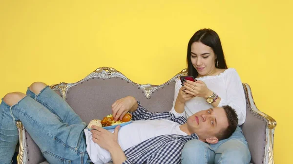 Chlápek a holka jedí rychlé občerstvení, chlap leží na dívčím klíně a dívka se dívá na telefon na žlutém pozadí. Koncept zdravé a nezdravé stravy. fast food — Stock fotografie
