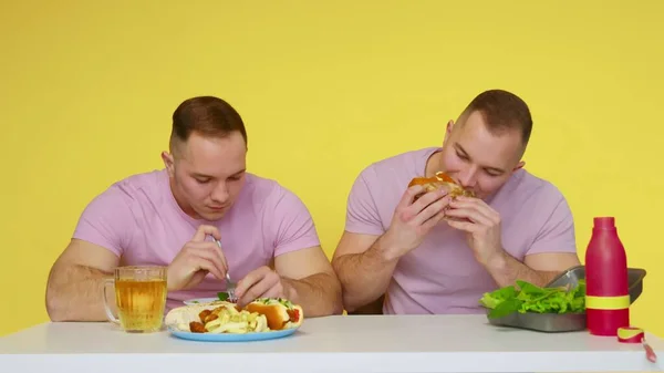 Két izmos iker egészséges ételt és gyorskaját eszik az asztalnál sárga alapon. Az egészséges és egészségtelen ételek fogalma. Gyorsétterem — Stock Fotó