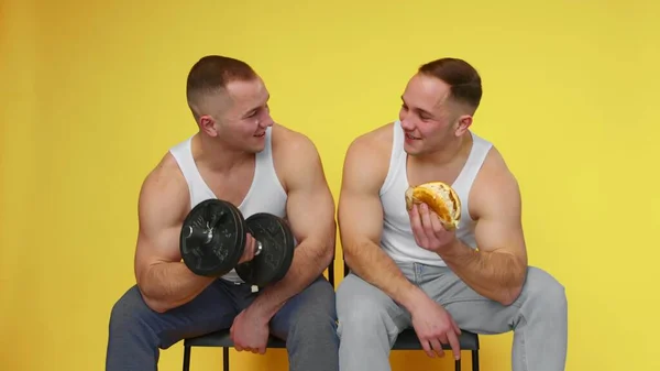 两个肌肉发达的双胞胎，背景是黄色的。一个人带着哑铃摆动，另一个人吃快餐。健康和不健康食品的概念。快餐食品 — 图库照片