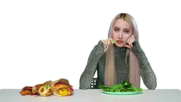 Egy aranyos lány zöldséget eszik, és szomorúan nézi a gyorskaját fehér alapon. Fogyókúra. Az egészséges és egészségtelen ételek fogalma. gyorsétterem — Stock Fotó