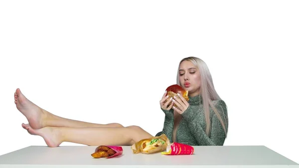 Симпатична дівчина сидить на стільці з ногами на столі і їсть гамбургер на білому тлі. Дієта. Концепція здорової та здорової їжі. фаст-фуд — стокове фото