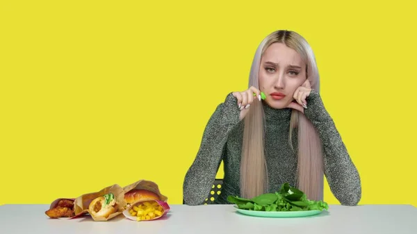 Ein nettes Mädchen isst Grünzeug und blickt traurig auf Fast Food auf gelbem Hintergrund. Ernährung. Das Konzept der gesunden und ungesunden Ernährung. fast food — Stockfoto