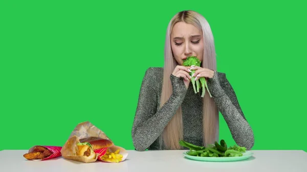Egy aranyos lány zöldséget eszik, és szomorúan nézi a gyorskaját zöld alapon. Fogyókúra. Az egészséges és egészségtelen ételek fogalma. gyorsétterem — Stock Fotó