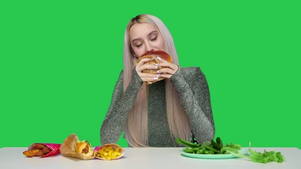 Uma garota bonita come verduras e olha tristemente para fast food em um fundo verde. A menina interrompe a dieta e come fast food. Dieta. O conceito de comida saudável e insalubre. fast food — Fotografia de Stock