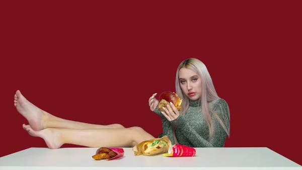 Egy aranyos lány ül egy széken, a lábaival az asztalon, és eszik egy hamburgert a piros háttér. Fogyókúra. Az egészséges és egészségtelen ételek fogalma. gyorsétterem — Stock Fotó