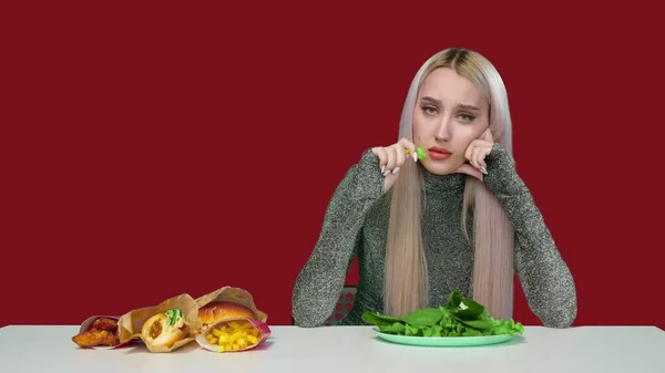 Una linda chica come verduras y tristemente mira la comida rápida sobre un fondo rojo. Dieta. El concepto de comida saludable y poco saludable. comida rápida — Foto de Stock