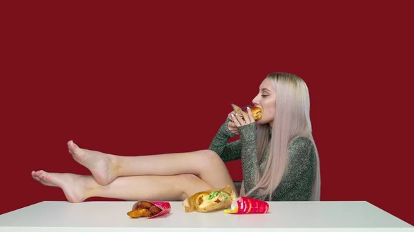 Egy aranyos lány ül egy széken, a lábaival az asztalon, és eszik egy hamburgert a piros háttér. Fogyókúra. Az egészséges és egészségtelen ételek fogalma. gyorsétterem — Stock Fotó