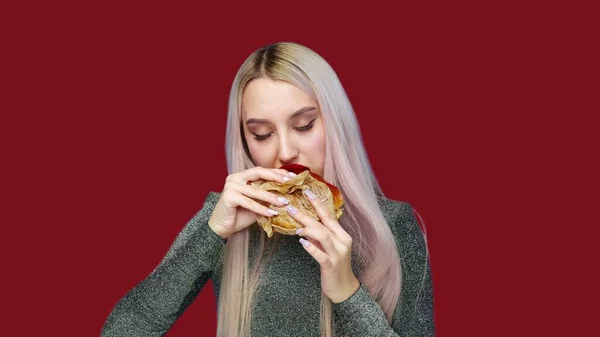 Close-up de uma menina com batom vermelho nos lábios, comendo um hambúrguer em um fundo vermelho. Dieta. O conceito de comida saudável e insalubre. fast food — Fotografia de Stock