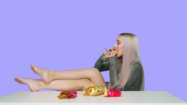 Egy aranyos lány ül egy széken, a lábaival az asztalon, és eszik egy hamburgert lila alapon. Fogyókúra. Az egészséges és egészségtelen ételek fogalma. gyorsétterem — Stock Fotó