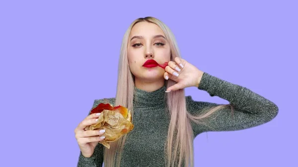 Közelkép egy lányról, piros rúzzsal az ajkain, hamburgert eszik és rúzst keneget a kezével a lila háttéren. Fogyókúra. Az egészséges és egészségtelen ételek fogalma. gyorsétterem — Stock Fotó