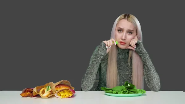 Мила дівчина їсть зелень і, на жаль, дивиться на фаст-фуди на сірому фоні. Дієта. Концепція здорової та здорової їжі. фаст-фуд — стокове фото