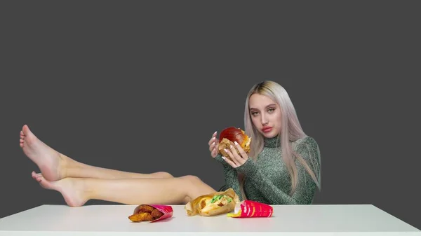 Egy aranyos lány ül egy széken, a lábaival az asztalon, és eszik egy hamburgert szürke alapon. Fogyókúra. Az egészséges és egészségtelen ételek fogalma. gyorsétterem — Stock Fotó