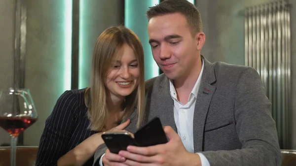 An attractive couple is sitting at a table using a smartphone, browsing the web, discussing online purchases, ordering food from a restaurant for a romantic dinner — Stock Photo, Image