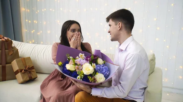 Um jovem dá flores à namorada enquanto está sentado no sofá. Romance. — Fotografia de Stock