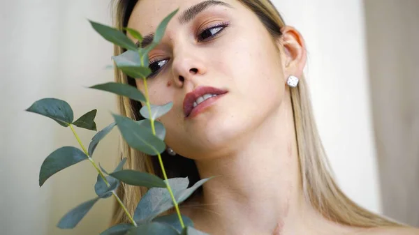 El rostro de una joven con la piel limpia y lisa y una pequeña rama en las manos. Maquillaje natural. La chica sostiene las hojas en su mejilla. Modelo posando para la cámara —  Fotos de Stock