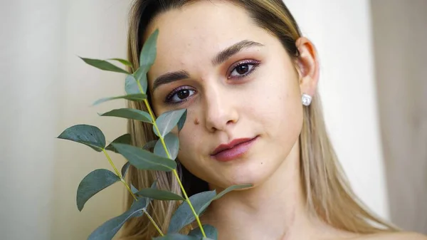 El rostro de una joven con la piel limpia y lisa y una pequeña rama en las manos. Maquillaje natural. La chica sostiene las hojas en su mejilla. Modelo posando para la cámara — Foto de Stock