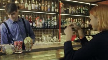Bir kız elinde bir kokteyl ile barda oturuyor ve barmenle iletişim kuruyor.