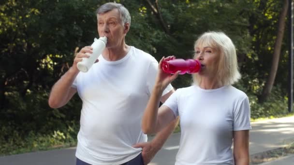 Koncepcja bilansu wody. Starszy mężczyzna i kobieta pijący z butelek sportowych, cieszący się przerwą w parku publicznym, śledzący strzał — Wideo stockowe