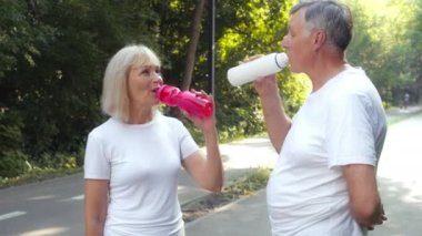 Bir çift aktif kıdemli adam ve kadın mola veriyor. Koşu antrenmanı yapıyor, şişelerden su içiyor, halka açık bir parkta duruyor.