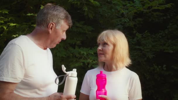 Mluvící starší muž a žena konverzace ve veřejném parku, stojící s lahvemi vody během ranního cvičení — Stock video