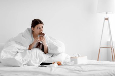 Üzgün, aç, beyaz, genç bir kadın evde oturmuş çikolata ve abur cubur yiyor.