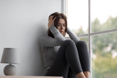 Depresyon, kriz ve stresten muzdarip üzgün, beyaz bir kadın elini tutup pencere eşiğinde oturuyor.