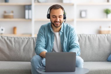 Kulaklıklı, gülümseyen beyaz bir adam. Evde bilgisayarla video görüşmesi yapıyor. Web kamerasıyla konuşuyor.