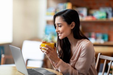 Genç bayan serbest çalışan kafede dizüstü bilgisayarla kahve içiyor, gülümsüyor ve bilgisayar ekranına bakıyor.