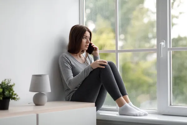 Khawatir tertekan kegelisahan Milenial perempuan duduk di jendela di rumah dan berbicara di telepon, menelepon untuk pacar — Stok Foto