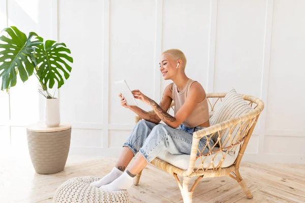 Κυρία που χρησιμοποιεί ψηφιακό tablet και ασύρματα ακουστικά, αναπαύεται σε ψάθινη καρέκλα πάνω από λευκό τοίχο, αντίγραφο χώρου — Φωτογραφία Αρχείου