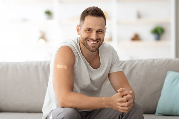 Joven hombre sano sentado en el sofá en casa con yeso médico en el brazo vacunado y sonriendo ampliamente a la cámara —  Fotos de Stock
