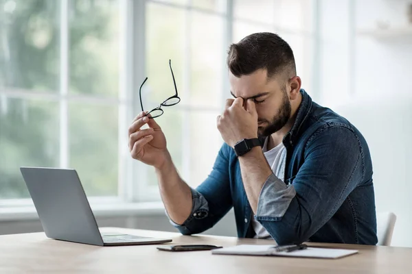 Разочарованный молодой человек в рубашке, с ноутбуком на удаленной работе, снимает очки, массирует глаза — стоковое фото