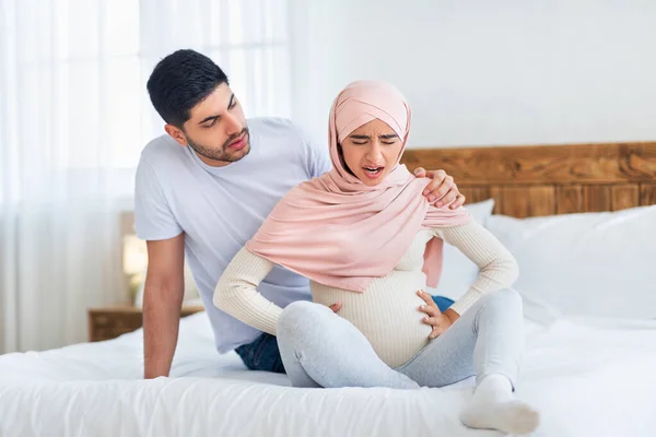 Bezorgde Arabische man ondersteunt zwangere vrouw met prenatale weeën, samen op bed zitten, kopieerruimte — Stockfoto