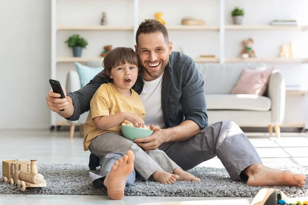 Passatempo familiar. Emocional jovem pai assistindo filmes na TV com seu filhinho, sentado no chão com pipocas — Fotografia de Stock