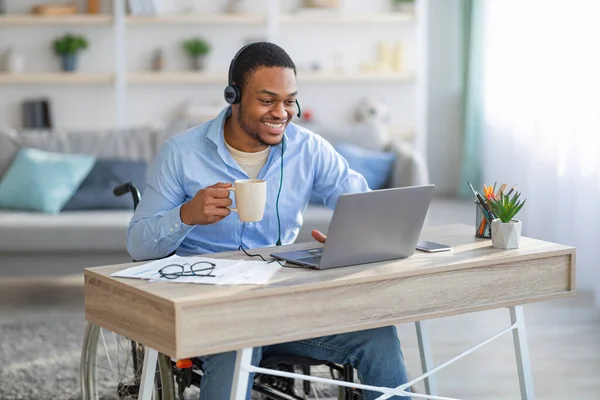 Engelliler için uzak bir iş. Tekerlekli sandalyedeki siyah adam kulaklık takıyor, laptop kullanıyor, ev ofisinde kahve içiyor. — Stok fotoğraf