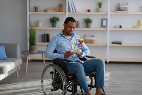 Boldogtalan fekete mozgássérült sportoló tolószékben trófeákkal a kezében, depresszióérzet otthon — Stock Fotó