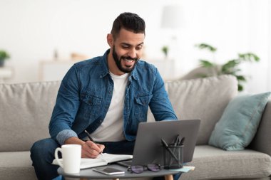 Bilgisayarda çalışan ya da ders çalışan mutlu genç bir Arap. Not alıyor, kapalı alanda iş görüşmesi yapıyor.