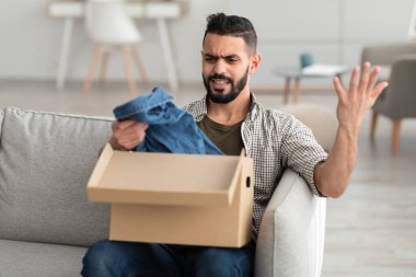 Hayal kırıklığına uğramış genç bir Arap karton paketi açıyor, giysiler alıyor, eve teslim edilen eşyalardan memnun değil.