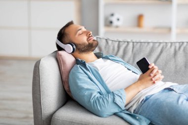Rahat genç adam, kulaklıkla kanepede uzanıyor, akıllı telefondan müzik dinliyor, yeni cep telefonu uygulaması kullanıyor,