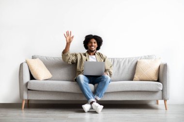 Neşeli siyah adam evde dizüstü bilgisayar kullanıyor ve kameraya el sallıyor.
