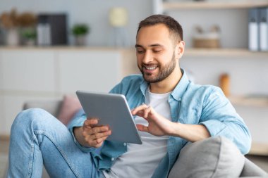 Tablet kullanan neşeli genç adam, evden internet üzerinden çalışıyor ya da çalışıyor