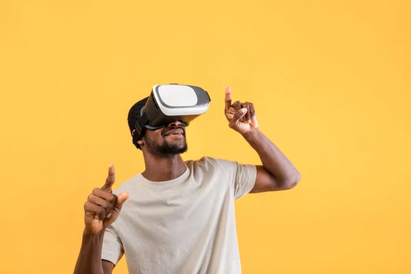 Afrykański Amerykanin w nowoczesnych okularach VR badający wirtualną rzeczywistość, poruszający dłońmi nad żółtym tłem — Zdjęcie stockowe
