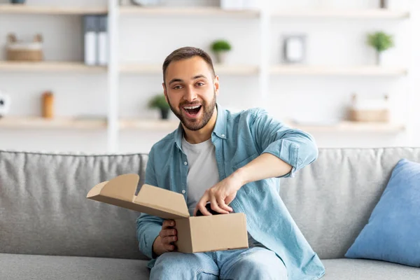 Портрет веселого молодого чоловіка, який отримує посилку, розпаковує пакет, виймає ігровий джойстик, сидить на дивані вдома — стокове фото